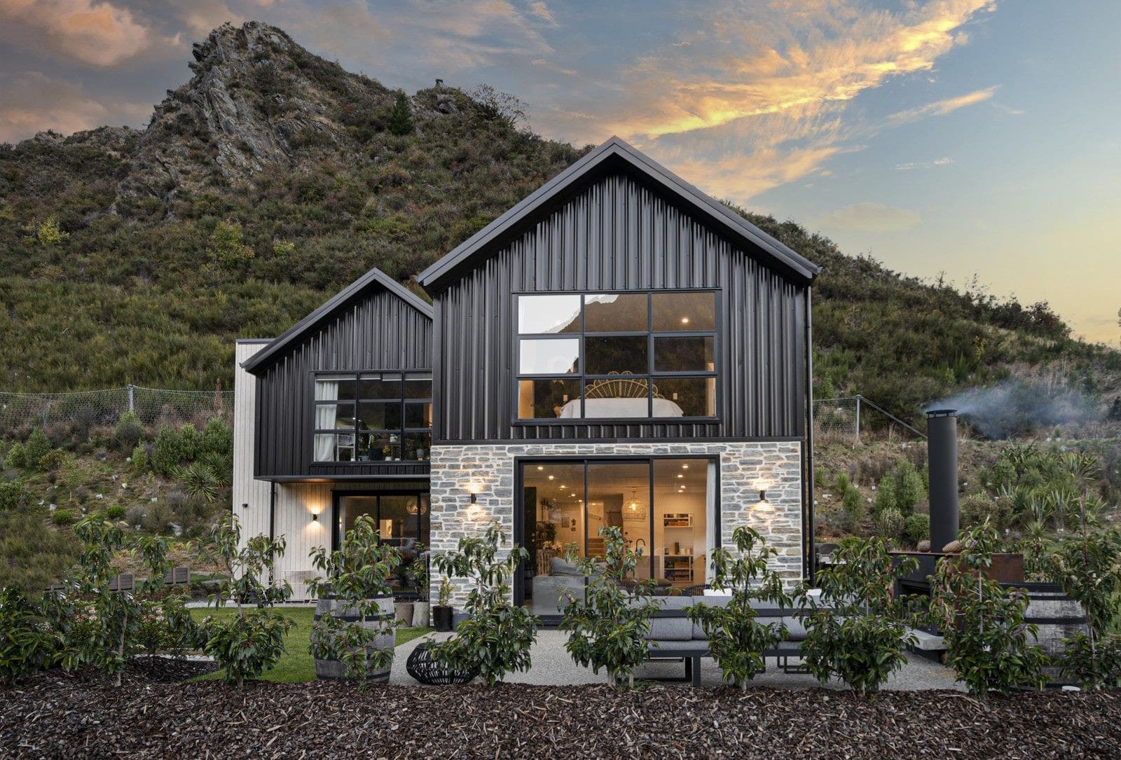 Gabled home in dark vertical cladding on a stone base, its windows lit at dusk below a rocky hillside
