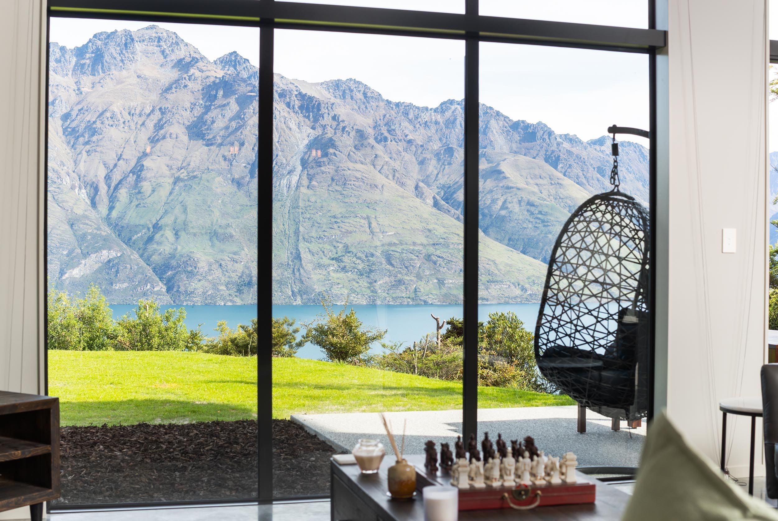 Full-height glazing framing the lake and the mountains, with a hanging chair beside it