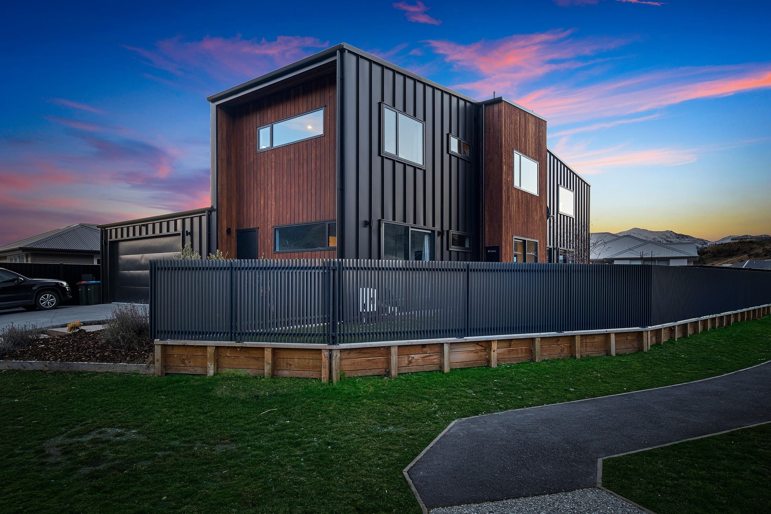 Two-storey home in dark vertical cladding with a timber-faced section, behind a dark fence at sunset